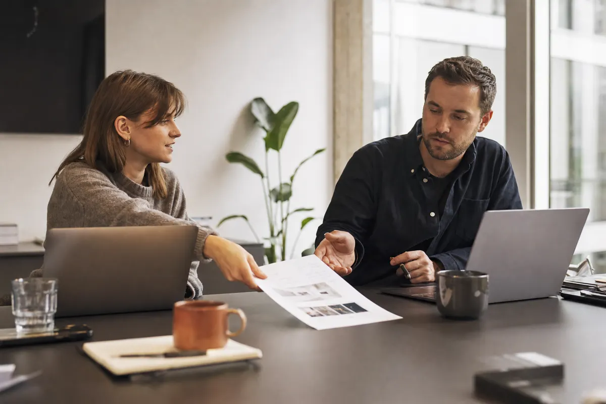 Two people at a meeting table handing over a printed project brief